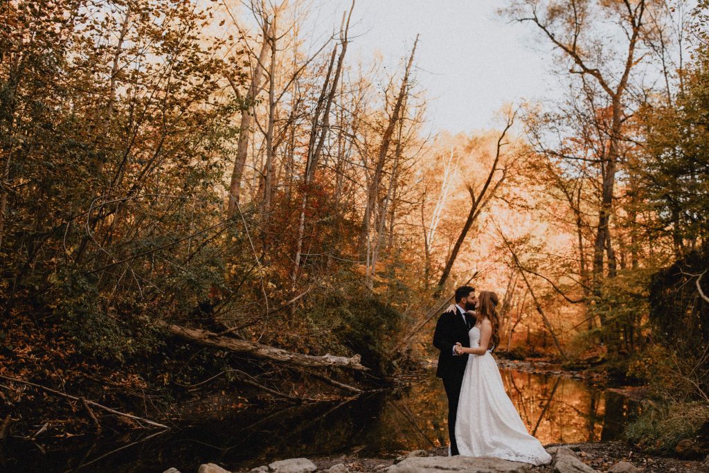 Bride and groom kiss beside river in back-lit forest - Autumn Micro Wedding at Berkeley Fieldhouse